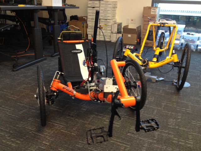 Two autonomous trikes, one orange and one yellow, sitting in front of a row of stacked boxes.