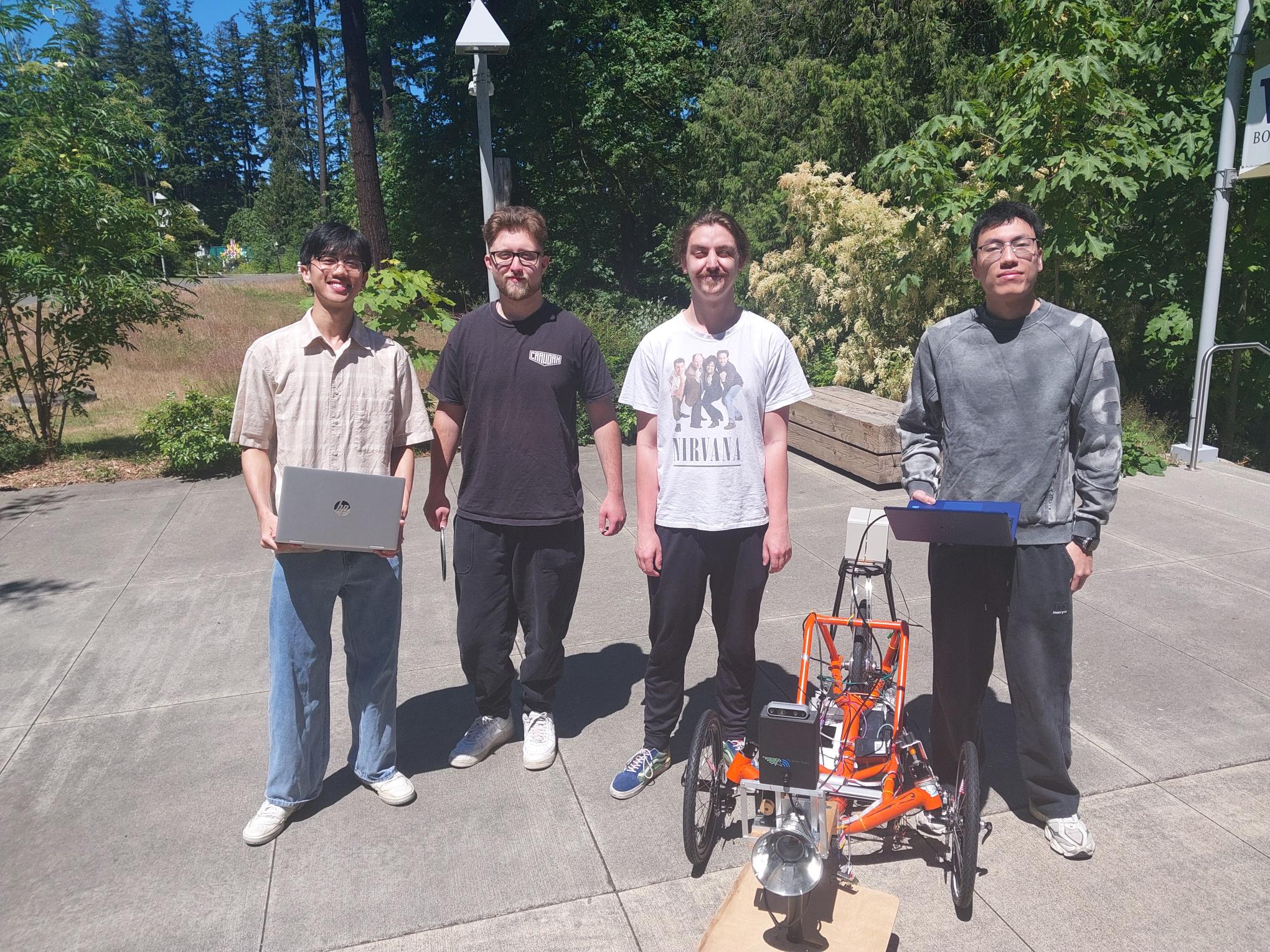 Four students stand outside with laptops behind an orange autonomous trike.