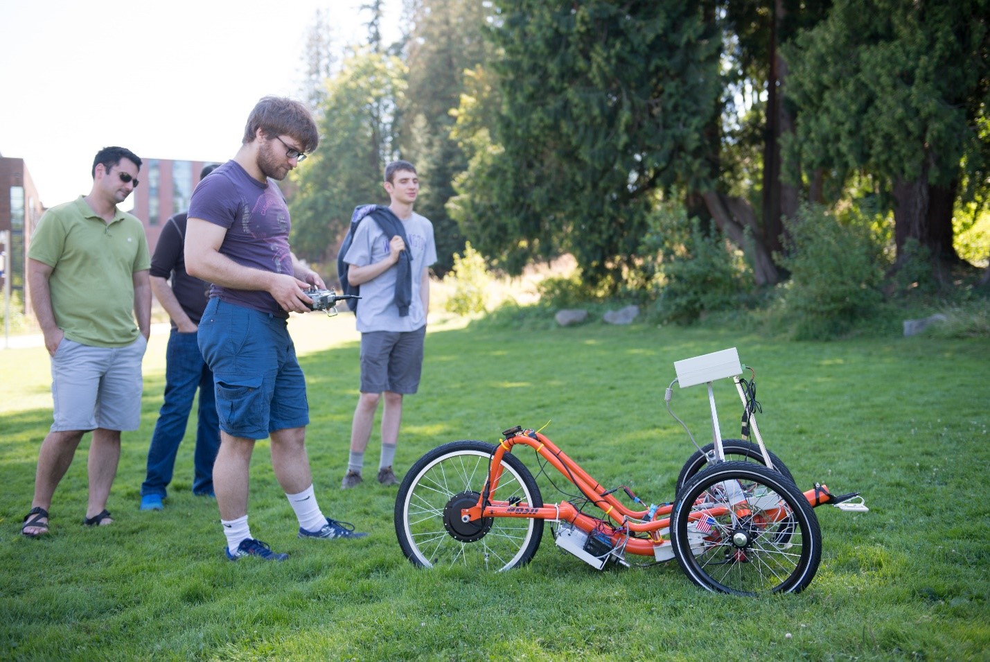 Four people stand behind an orange autonomous trike in a grassy field.
