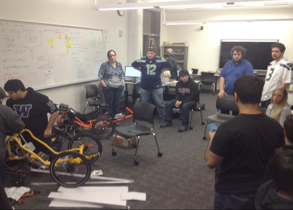 A classroom of students surrounding two autonomous trikes, one orange and one yellow. There is a whiteboard and many chairs.