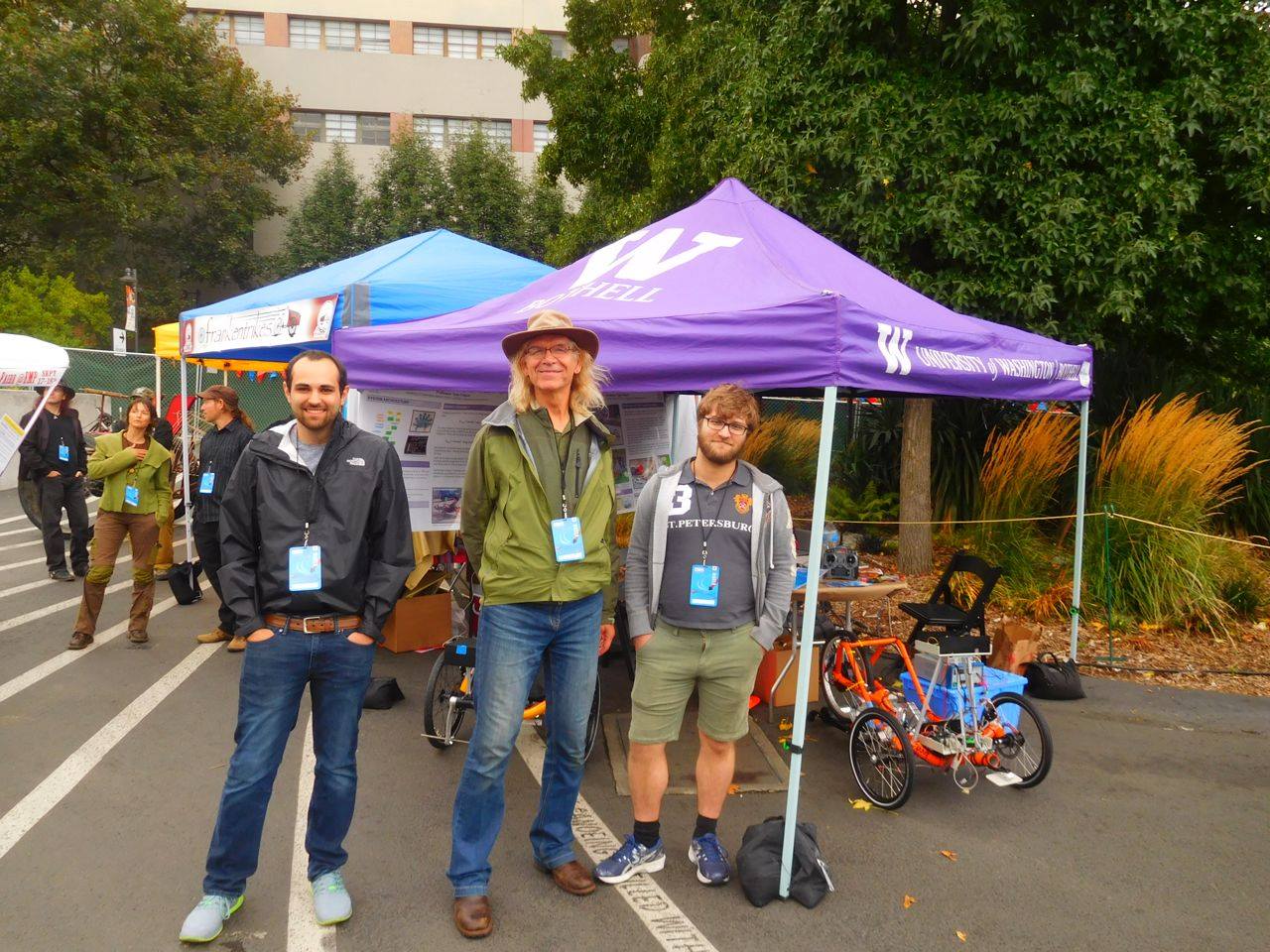 Three people stand with an orange autonomous trike in front of a purple canopy tent.