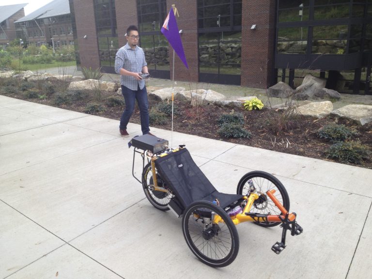 A person remote controls a yellow autonomous trike on a paved trail.