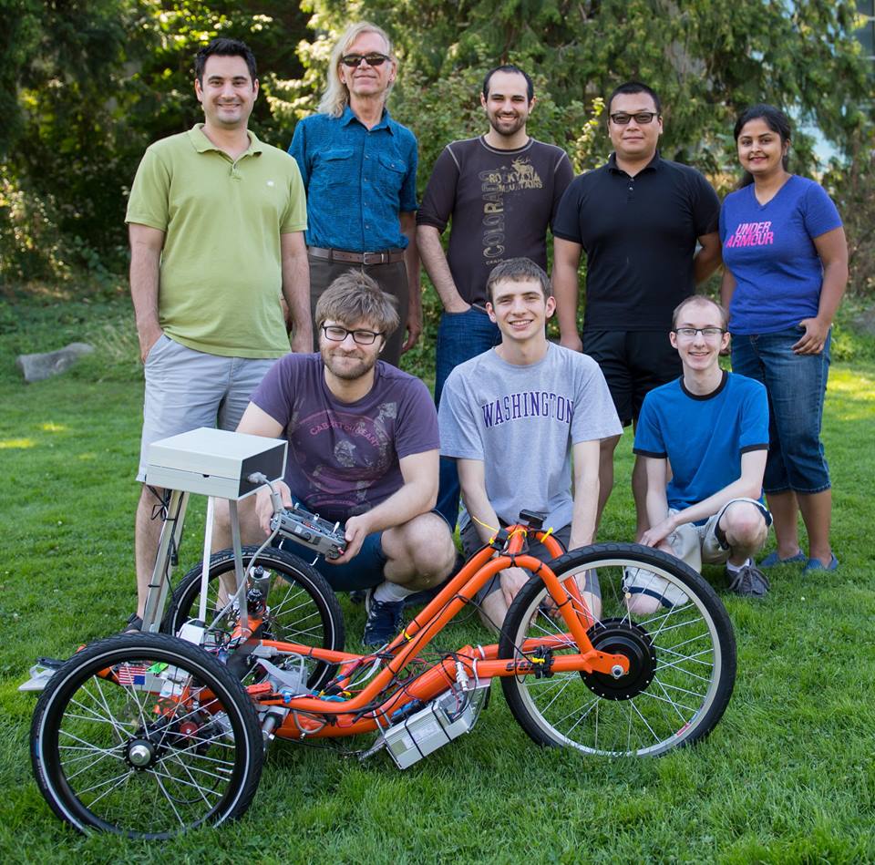 Five people stand and three people kneel behind an orange autonomous trike in a grassy field in front of trees.