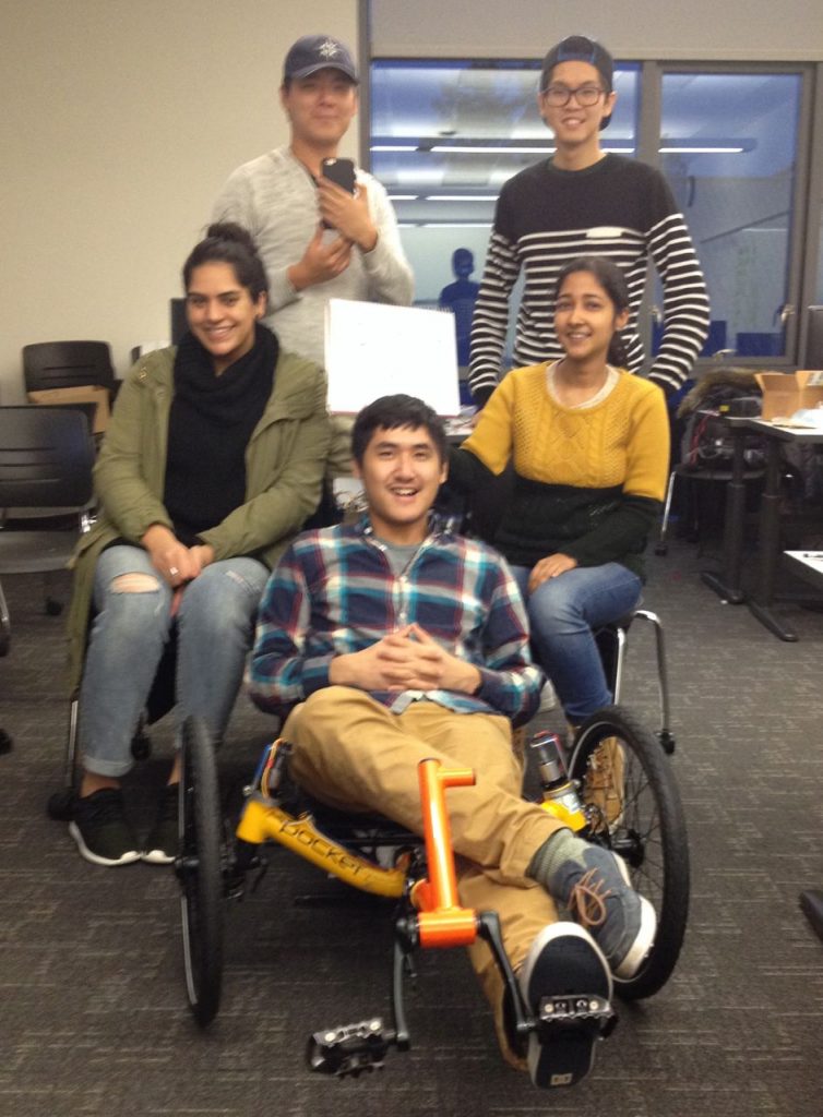 Five people smile around a yellow autonomous trike in a classroom.