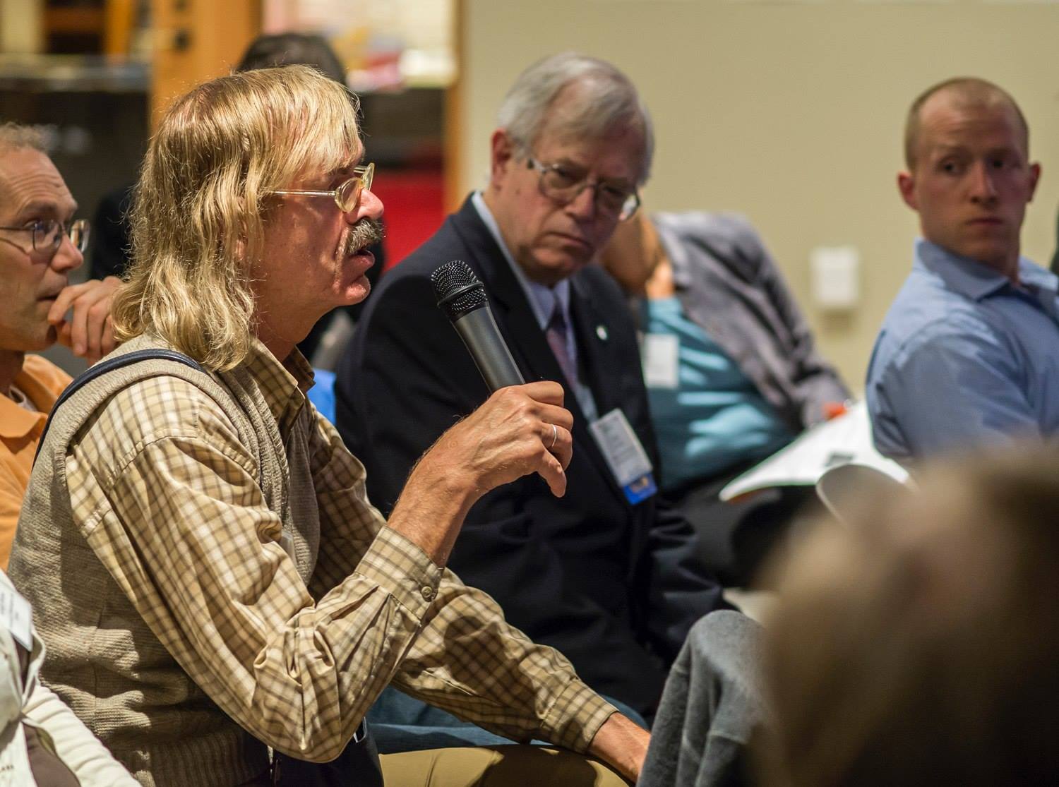 A person sitting down in a brown shirt speaks into a microphone. Others listen attentively.