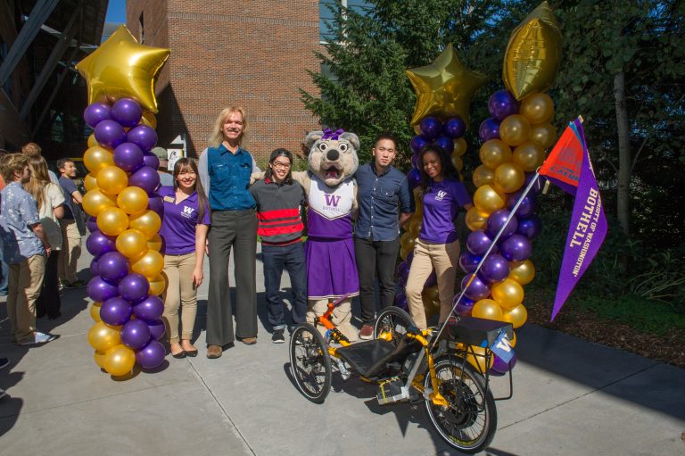 Five people and a Husky mascot stand between purple and gold balloon columns behind a yellow autonomous trike.